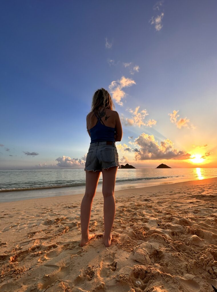 A blonde woman in a blue shirt and jean shorts looking towards the ocean sun rising over islands